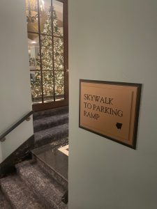 Skywalk to Parking Ramp Sign with Festive Decor A wall-mounted sign indicating the direction to the skywalk leading to the parking ramp, with a festive Christmas tree visible through a glass window in the background.