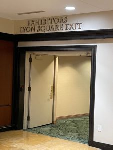 Exhibitors Lyon Square Exit An interior hallway exit labeled "EXHIBITORS LYON SQUARE EXIT" with an open white door leading to a carpeted corridor, framed by dark wood trim.