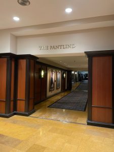 The Pantlind Hallway Entrance A hallway entrance labeled "The Pantlind," featuring classic wood paneling, framed portraits, decorative wall sconces, and a carpeted walkway leading further inside.