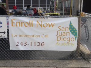 San Juan Diego Academy Enrollment Banner A promotional banner for San Juan Diego Academy, encouraging enrollment with a contact phone number 243-1126, displayed on a chain-link fence.