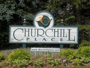 Churchill Place Entrance Sign A rustic entrance sign for Churchill Place featuring a wood-textured background, bold green lettering, a decorative French horn emblem, and a website address "www.churchillplace.com," surrounded by landscaped greenery.