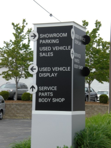 Automotive Dealership Directional Sign A black and white directional sign at an automotive dealership, guiding visitors to showroom parking, used vehicle sales, display, service, parts, and the body shop.