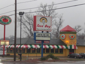 Taco Boy Restaurant Sign and Fazoli's in the Background A Taco Boy restaurant sign with the slogan "Home of the Super Burrito," advertising a 99-cent Monday menu deal, with a Fazoli's restaurant in the background featuring a red, white, and green awning.