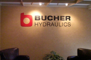 Bucher Hydraulics Office Wall Sign An interior office wall featuring the Bucher Hydraulics logo with a red and white emblem and bold black text, flanked by two black leather chairs and a decorative plant.