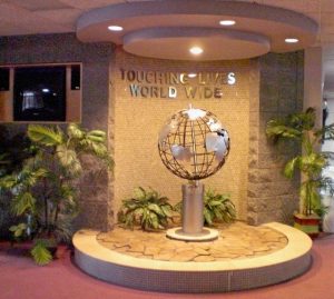 Touching Lives Worldwide Globe Display An indoor decorative display featuring a metallic globe sculpture on a pedestal with the phrase "Touching Lives Worldwide" mounted on a tiled wall, surrounded by greenery.
