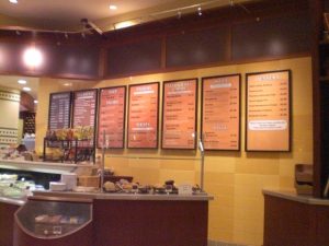 Cafe Restaurant Menu Board A cafe-style restaurant interior featuring an illuminated menu board displaying categories such as beverages, breakfast, soups, salads, paninis, sandwiches, pizza, pasta, and desserts.